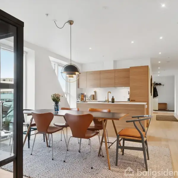 Et moderne køkken-alrum med et rundt spisebord omgivet af brune stole. Over bordet hænger en sort pendel. I baggrunden ses et lyst køkken med træskabe og en åben altandør.