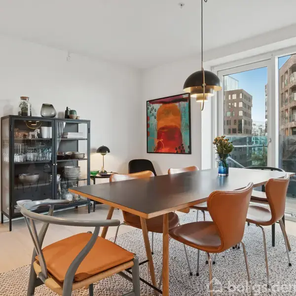 En spisestue indeholder et træbord med seks stole. På bordet står en vase med blomster. Et stort vindue viser en byudsigt. En metalreol og maleri er bagved.