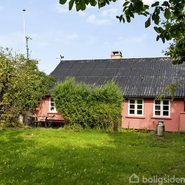 Et lyserødt hus med sort tag står i en grøn have med frodige træer og buske omkring. Ingen tekst fundet.