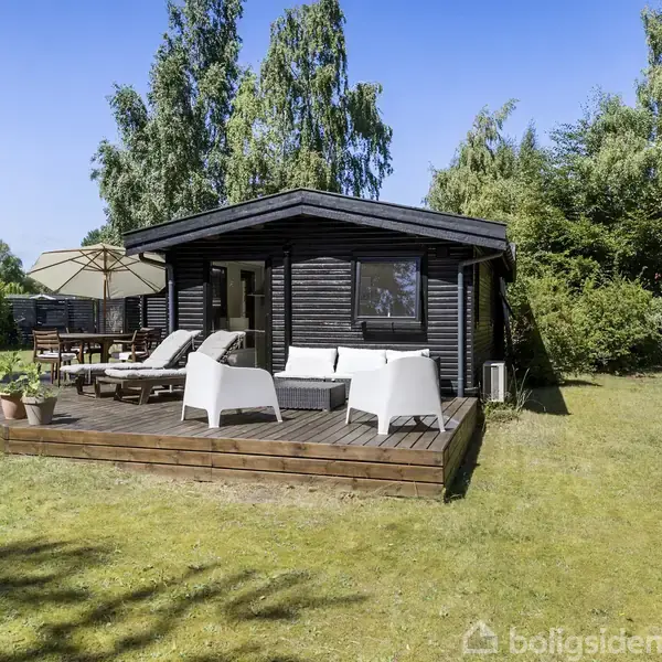 Sort træhus med terrasse, hvor der er placeret hvide møbler og urtepotter. Terrassen er omgivet af frodig have med høje træer og græsplæne.
