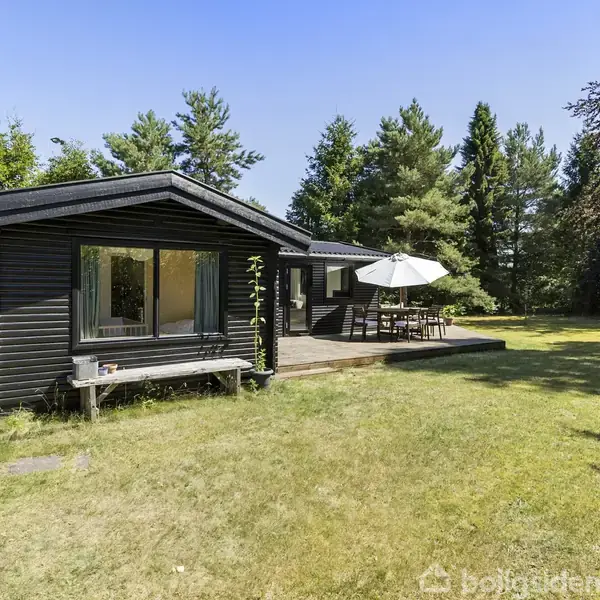 Sort træhus står stille omgivet af grønne træer og græsplæne. Ved siden af huset er en terrasse med et bord og en parasol, der skaber en hyggelig udendørs atmosfære.