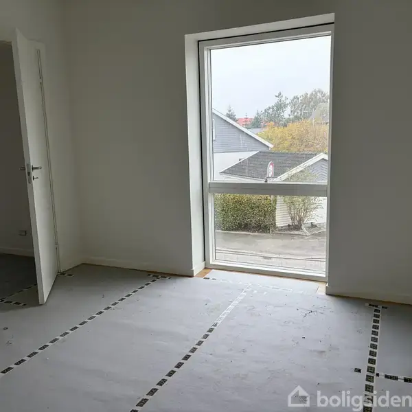 Et tomt rum har et stort, rektangulært vindue. Gulvet er dækket med beskyttende papir. En åben dør til venstre viser en tilstødende gang. Udenfor ses huse og træer.
