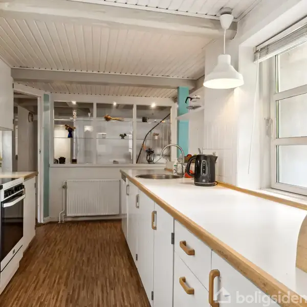 Et lyst køkken med trælaminatgulve har hvide skabe og bordplader. En elkedel står ved vasken under et stort vindue. Lyse vægge og loftslampe skaber en indbydende atmosfære.