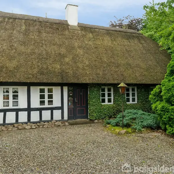 Et bindingsværkshus med stråtag står stille omgivet af grønne buske og træer. Foran huset er der en grusbelagt gårdsplads.