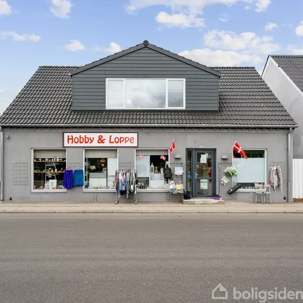 Butik Hobby & Loppe viser varer udenfor, herunder tøj på stativer og danske flag. Bygningen er grå med et mørkt tag, beliggende mellem andre huse på en gade.