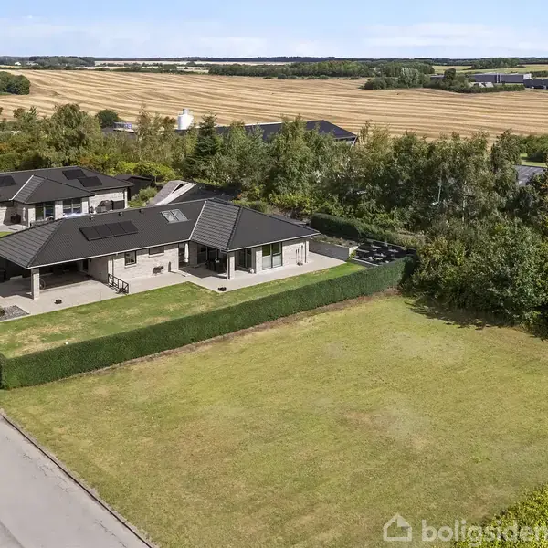 Et moderne hus med sort tag ligger ved siden af en grøn hæk i et åbent, landligt landskab med flere huse og marker i baggrunden under en blå himmel.