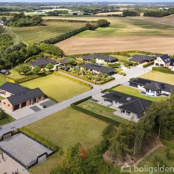 Et parcelhuskvarter med moderne huse og velplejede haver. I forgrunden ses en legeplads med gynger. Området er omgivet af marker og skovområder i baggrunden.