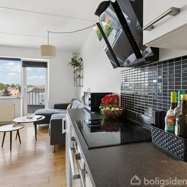 Et køkken-alrum med kogeplader og sort køkkenbord. I baggrunden er der en sofa og runde borde. Stort vindue med udsigt til træer og skyer i fjerne horisont.