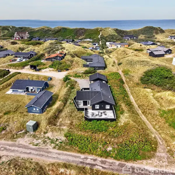 Sort sommerhus med terrasse omgivet af græs og sandklitter, placeret i et naturskønt område med flere andre huse. Baggrunden viser et smukt, bjergrigt landskab ved kysten.