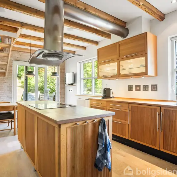 Et moderne køkken med træskabe og en ø med indbygget kogeplade, under en emhætte. En trappe fører til en anden etage. Store vinduer giver lysindfald.
