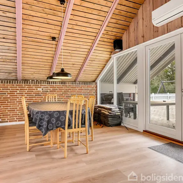 Et spisebord med fire træstole står stille på en trælaminatgulv. Det er dækket med en sort dug. Omkring er der en skråt loft med brædder og glasdøre til en terrasse.