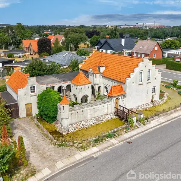 Et hvidt hus med rødt tegltag har en slotlignende arkitektur med buer og tårne. Huset ligger på en hjørnegrund omgivet af veje og grønne haver i et beboelseskvarter.