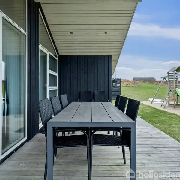 Udendørs bord med otte sorte stole på terrasse foran moderne sort hus. I baggrunden ses en græsplæne med en legeplads og klart, blåt himmel.