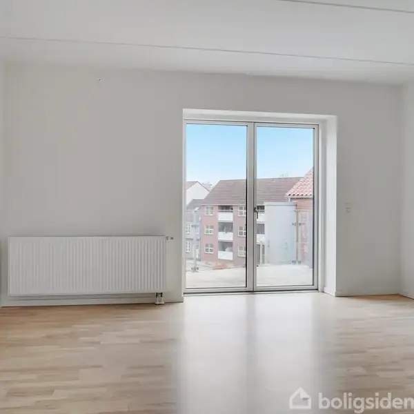 Et lyst rum med trægulv og en radiator, hvor store glasdøre åbner ud til en balkon med udsigt over omkringliggende bygninger. Ingen tekst er synlig.