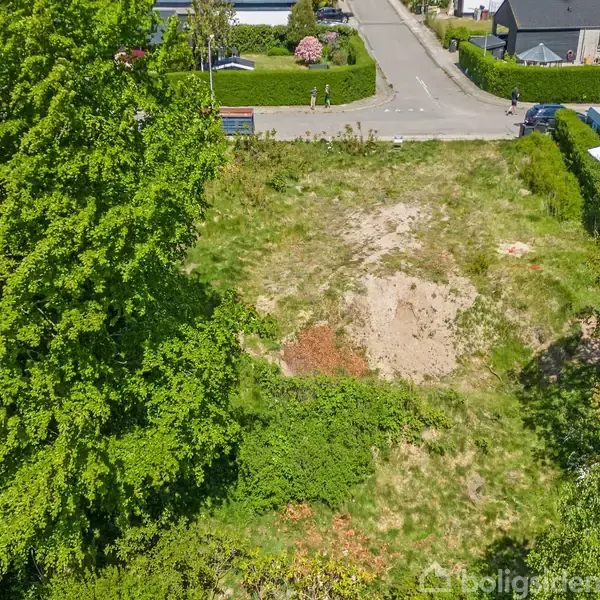 Ledigt byggegrund med ujævne jordpletter omgivet af grønne træer og buske. Grunden ligger i et boligområde med veje og huse synlige i baggrunden.