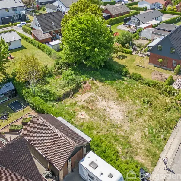 Et tomt græsareal ligger mellem to huse. Omkring arealet er der andre huse og græsplæner. Fortovet løber langs siden af grunden i et beboelseskvarter.