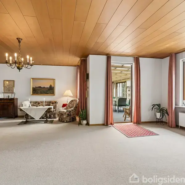 En rummelig stue med tæppebelagt gulv, træloft og en lysekrone. En sofa og bord står ved en lampe, mens store vinduer og glasdøre giver udsigt til en veranda og frodige omgivelser.