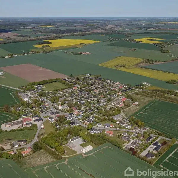 By fra luften, omgivet af store grønne og gule marker, med huse spredt blandt træer. Landskabet er fladt og strækker sig mod horisonten under en klar blå himmel.