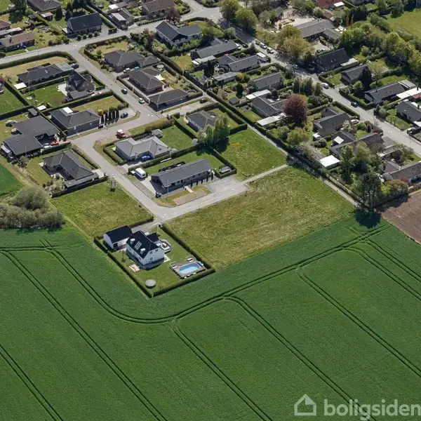 Hus med pool ligger i et fredeligt boligområde omgivet af marker og andre boliger. Husene er arrangeret i rækker med veje imellem, og markerne viser grønne afgrøder.