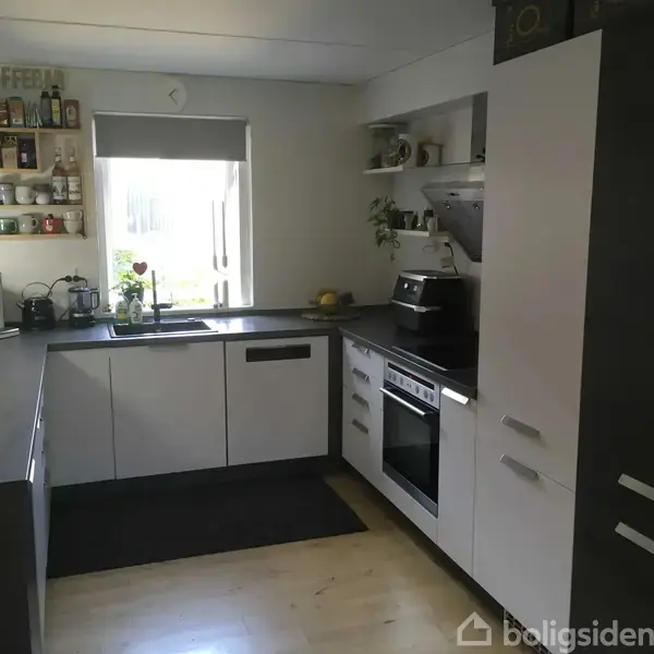 Køkkenet har hvide skabe og sorte bordplader, med en ovn og vask. Der er hylder med krydderier og flasker ved vinduet, hvor lyset kommer ind.
