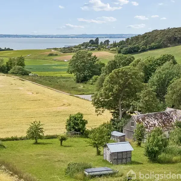 En gammel lade står stille i et frodigt landskab med grønne marker og en skovklædt bakke i baggrunden. En fjord strækker sig ud i horisonten under en klar himmel.
