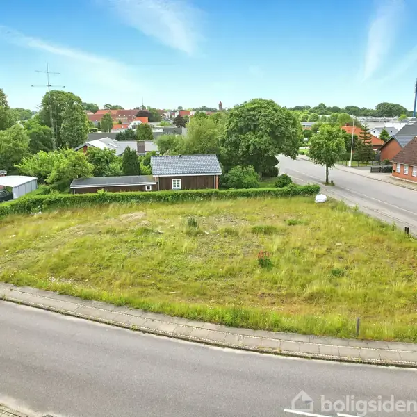 En græsbevokset tom grund ligger i en by med spredte huse og træer i baggrunden. Vejen omkranser grunden, og små gader er synlige i nærheden.