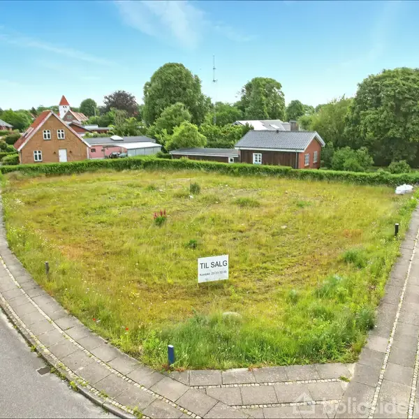 Grunden er dækket af græs med et skilt, der siger "TIL SALG". Den ligger i et villakvarter med træer og huse rundt om.