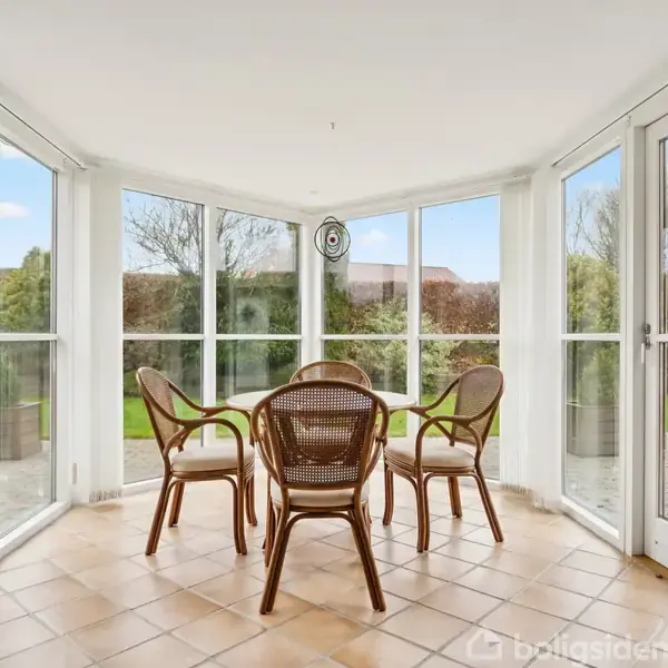 Fire flettede stole står omkring et rundt bord i en lys vinterhave med flisegulv, omgivet af glasvægge. Udenfor ses græsplæne og træer under en blå himmel.