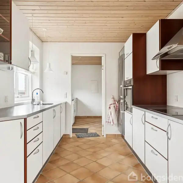 Køkken med hvide skabe og stålbordplader, der løber langs begge sider. En vask er placeret under et vindue til venstre. Terrazzogulv og træloft skaber en varm atmosfære.