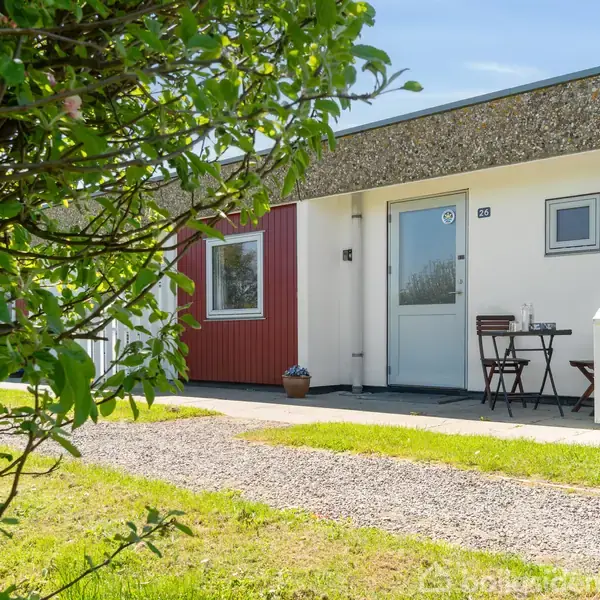 En lille terrasse foran et moderne, rødt og hvidt hus med et bord og to stole. Grussti og græsplæne omgiver huset. Tallet "21" ses ved døren.
