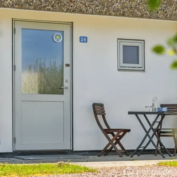 Dør markeret med nummer 26 står ved siden af et lille bord med to stole. Bordet har genstande på, inklusiv glas. Omgivet af en hvid mur og grønne planter.