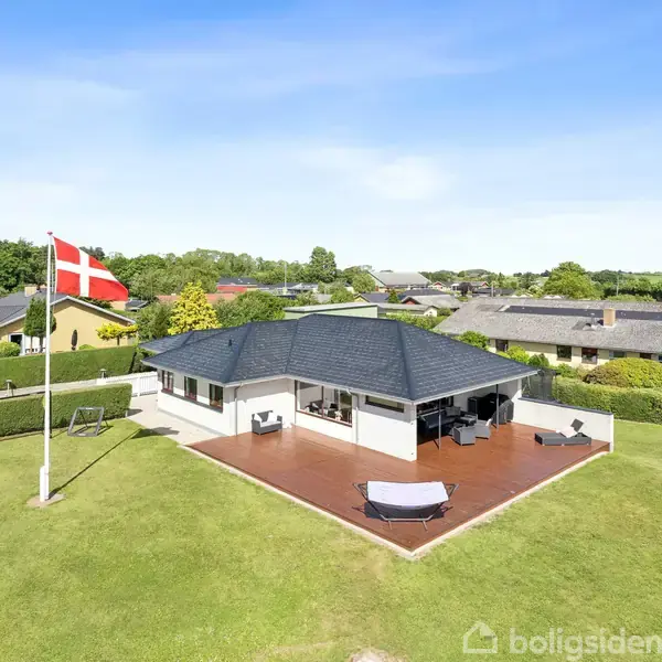 Et moderne hus med fladt, mørkt tag står i en velplejet have. En dansk flagstang står til venstre, og terrasseområdet omgiver huset. Omgivet af hække i et boligområde.