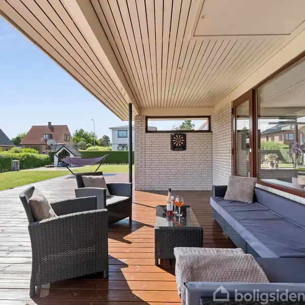 Terrasse med grå rattanmøbler står stille under et overdække. En dartskive hænger på husets mur, omgivet af grønne haver og nærliggende huse i baggrunden under en klar blå himmel.
