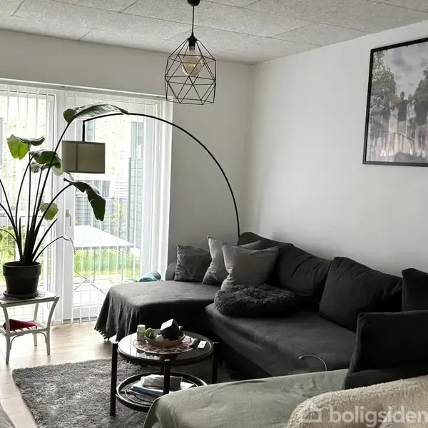 En stor, grå sofa står i en lys stue med en buet gulvlampe ved siden af. Foran er der et rundt sofabord, og en stor plante står ved siden af vinduet med persienner.