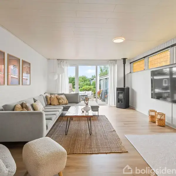 Stue med grå sofa og to stole, hvorfra man kan se et stort TV på væggen. Rummet har lys indretning og store vinduer mod en terrasse udenfor.