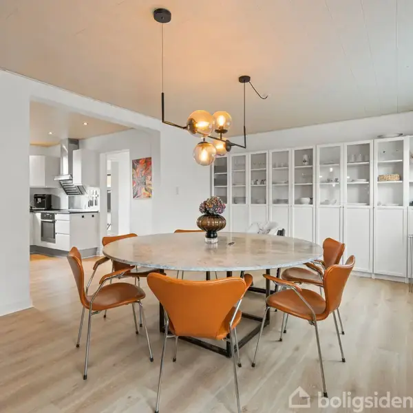 En rund spisebord omgivet af seks brune stole står i en lys spisestue med trægulv, moderne loftslampe, hvidt vitrineskab og vindue. I baggrunden ses et åbent køkken.