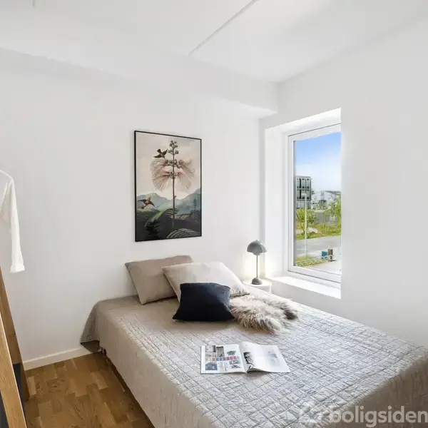 En seng med gråt sengetæppe ligger ved et vindue med udsigt over grønt område. På sengen ligger puder, en pelsdækken og et åbent blad. Et minimaliskt kunstværk hænger på væggen.