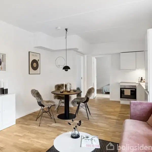 Sofa i lyserød står i lys moderne stue med træparket. Rundt bord omgivet af stole under pendellampe. Køkken ses i baggrunden gennem åben dør. Indrammet kunst på væggene.