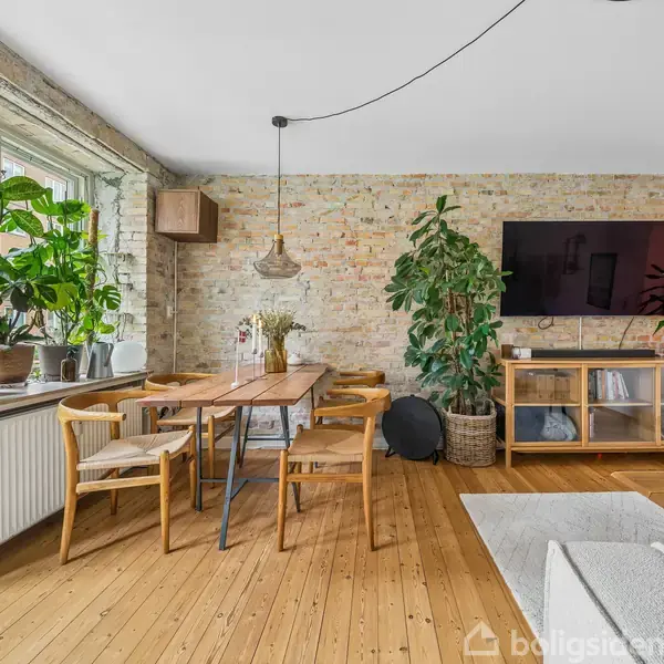 Spisestue med træbord og stole, placeret ved et vindue med flere grønne planter. Til højre ses en væg med TV og skænk. Rummet har lyse trægulve og rå murstensvægge.