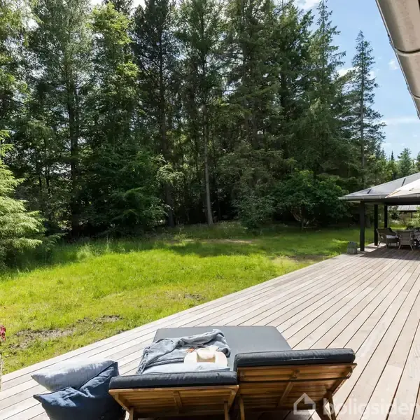 Terrasse med træstole og puder ved siden af et hus. En grøn græsplæne og tæt skov i baggrunden skaber en fredfyldt atmosfære i det åbne udemiljø.