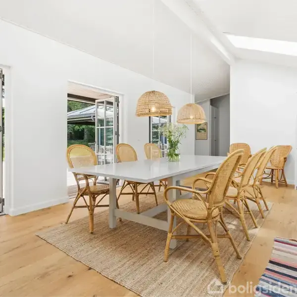 Et lyst, moderne spisekøkken med et langt bord omgivet af syv flettede stole. Store glasdøre åbner ud til en terrasse med grøn udsigt, mens loftslamper hænger over bordet.