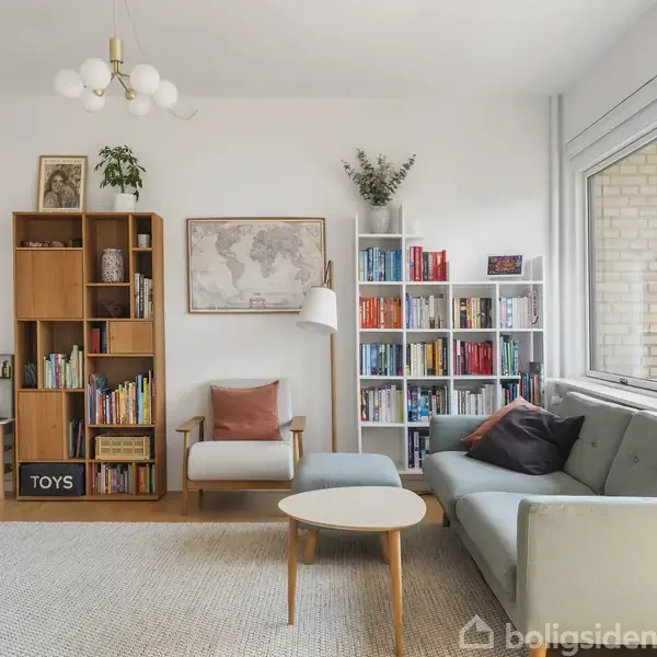 Stue med sofa og lænestol ved et sofabord. Hylder fyldt med bøger og dekorative genstande. Vægkort og planter hænger over lænestolen. Store vinduer lyser rummet op.