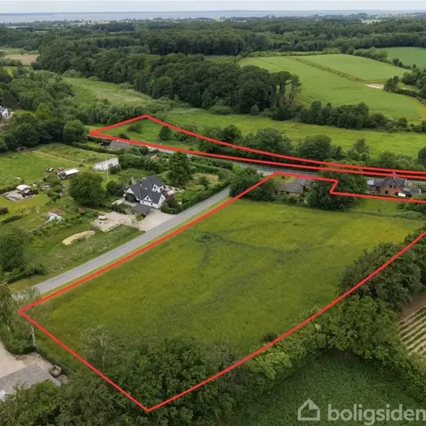 Landområde markeret med røde linjer strækker sig over grønne marker og et vejnet; omgivet af skove og enkelte huse i landligt område under en overskyet himmel.