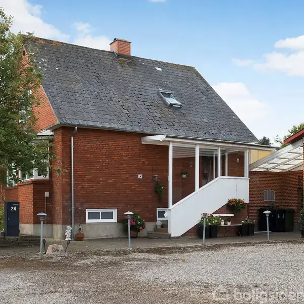 Et rødt murstenshus med en trappe op til en tildækket veranda. Foran er en grusbelagt indkørsel med blomsterkrukker. Til venstre står et træ. Husnummeret er 24.