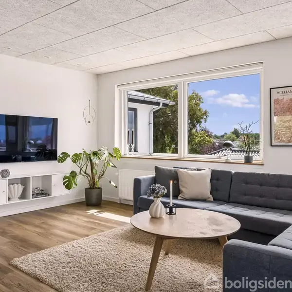 En moderne stue med en grå hjørnesofa og et rundt træbord. Et TV hænger på væggen. Store vinduer slipper naturligt lys ind, og en grøn plante står i hjørnet. Plakat på væggen.