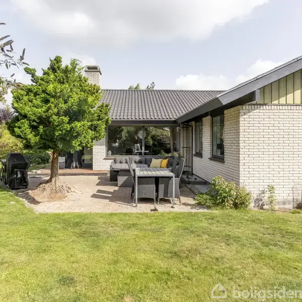 Huset er et énplans murstensbyggeri med et sort tag. Der står havemøbler og en grill på en terrasse af fliser, omgivet af en græsplæne, træer og buske.