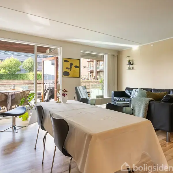 En moderne stue med et spisebord dækket med en hvid dug. I baggrunden er der en sofagruppe. Store vinduer og en glasdør fører ud til en terrasse med stole og bord.