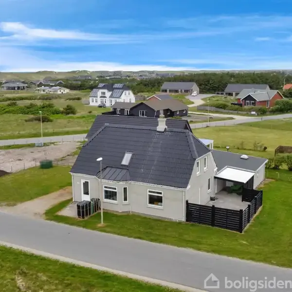 Hus set fra luften med mørkt tag, omgivet af en græsplæne. Ligger i et villakvarter med flere huse omkring og en vej foran. Lyseblå himmel og spredte skyer.