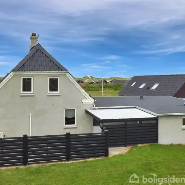Et hvidt hus med sort tag står på en græsplæne omgivet af lavt sort stakit. I baggrunden ses flere huse og blå himmel med spredte skyer.