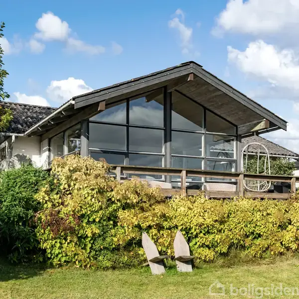 Et moderne hus med store glaspartier står omkranset af frodig vegetation i en have under en blå himmel med skyer. En terrasse med hængestol og parasol ses foran.